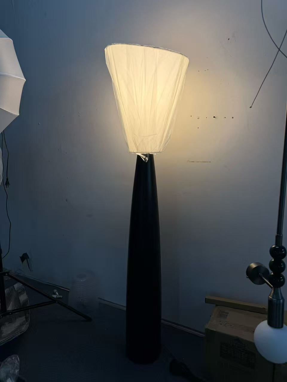 Tall floor light with a slender black tapered body and softly illuminated flared shade in the workshop