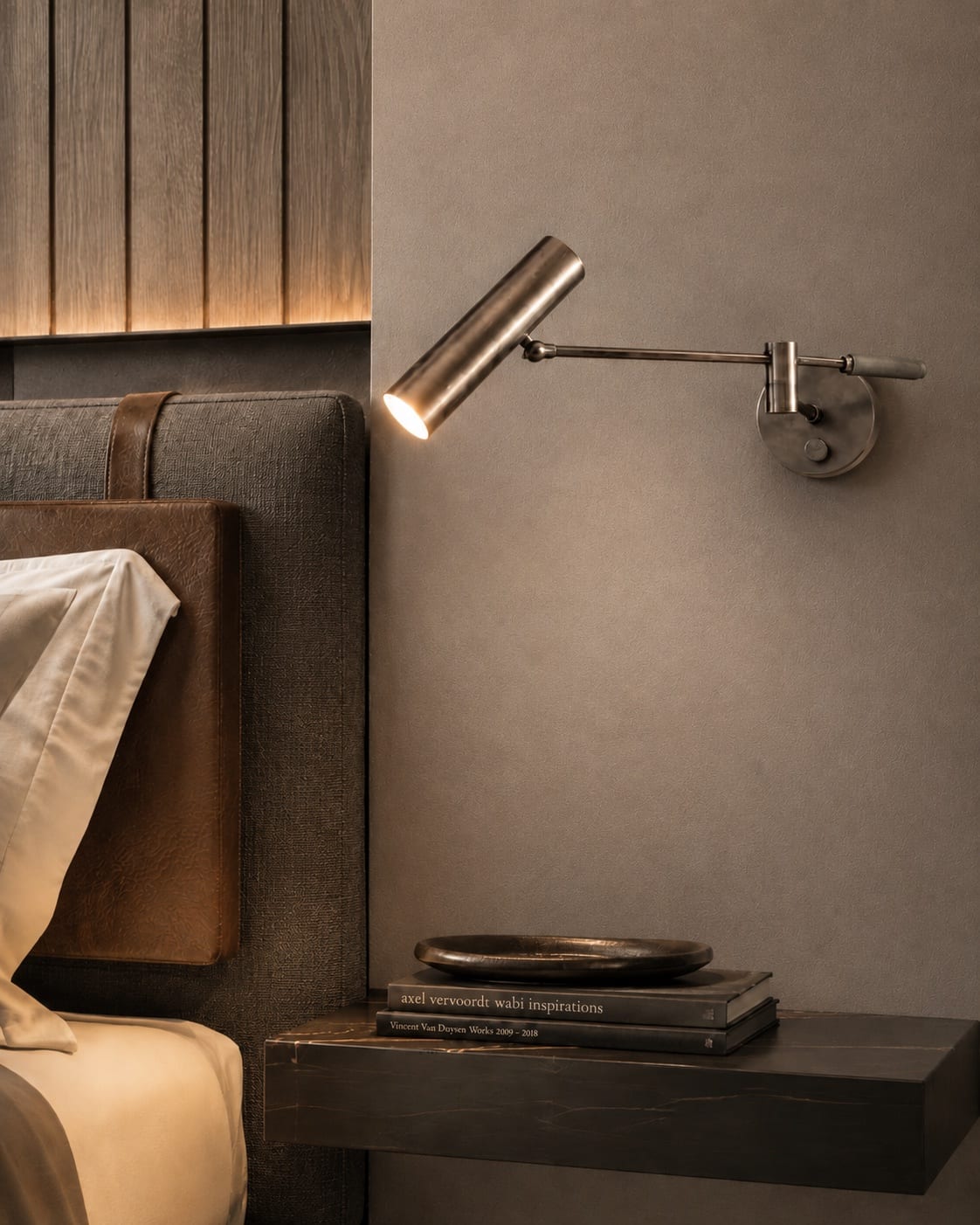 Articulated bronze bedside reading light extending from a circular wall plate in a warm bedroom interior